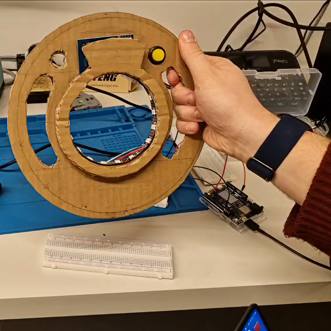 A hand holding a cardboard Curio with a yellow button wired to a breadboard