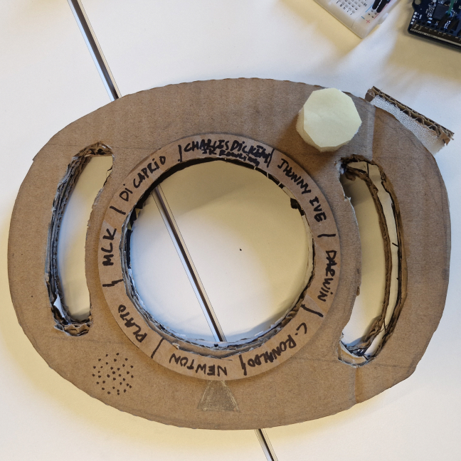 A cardboard ring with handles and a bottle-cap button, names written around the lens